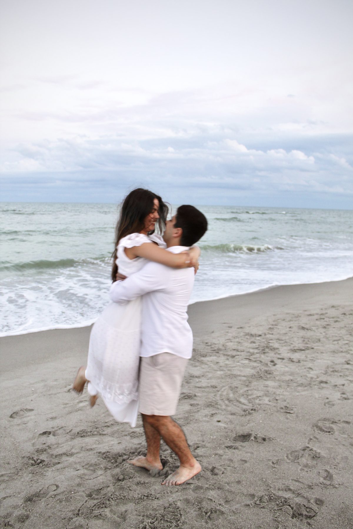 Couple in white embracing at the water's edge, he lifts her just off the sand