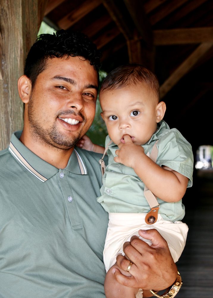 Dad in a sage-green polo holding his baby boy, who looks curiously at the camera