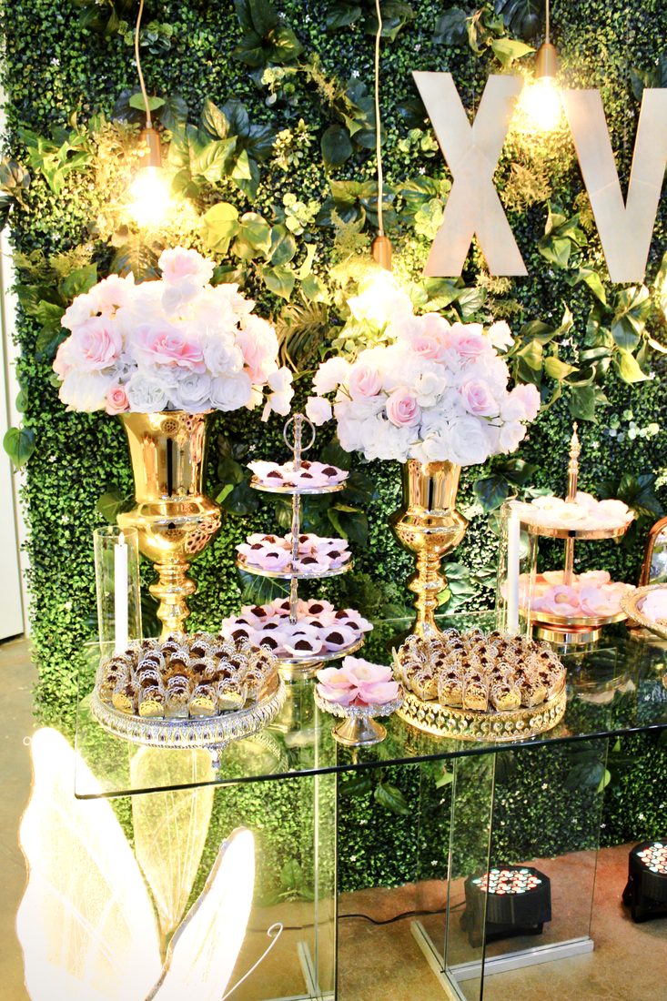 Dessert table with gold stands, pink flowers and treats in front of a floral wall with a large XV sign