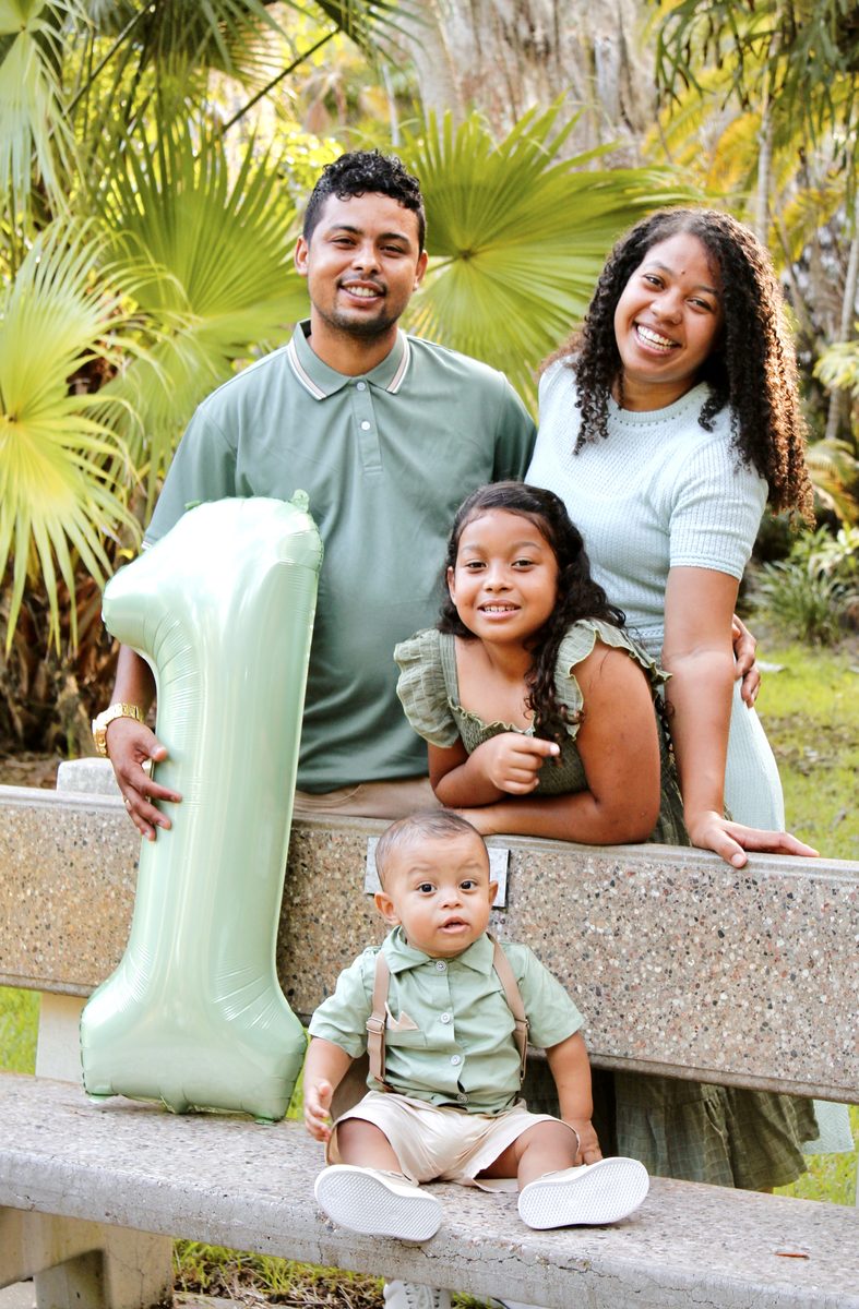 Family of four in matching sage green with their baby and a giant balloon number 1 on a stone bench