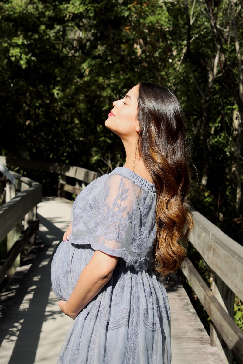 Expecting mother in a gray-blue dress on a wooden boardwalk, her face lifted to the light