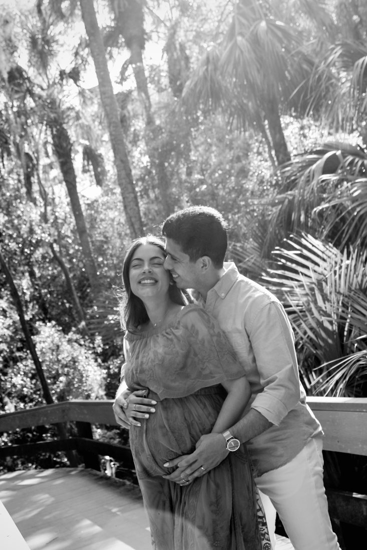Black-and-white photo of an expecting couple embracing among palms, his hands on her belly