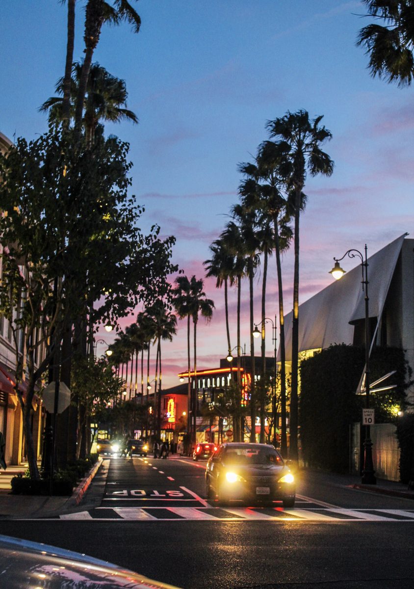 Palm-lined street at dusk with glowing lights, a pink and purple sky and a car approaching