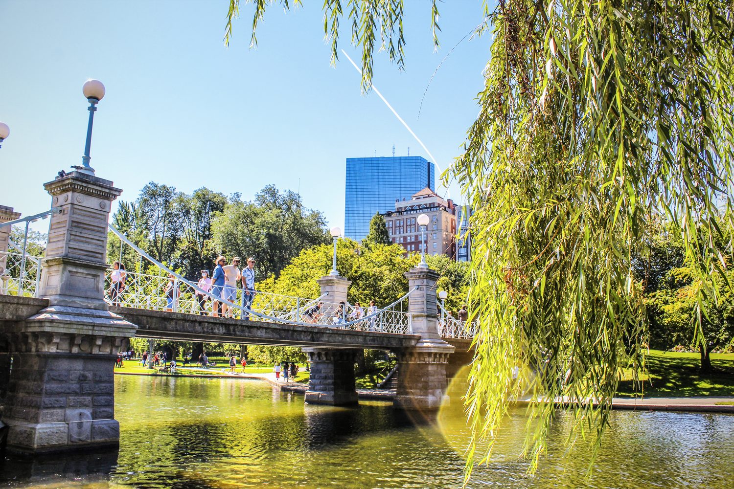 Stone bridge over a pond with a weeping willow and city buildings behind, under a blue sky