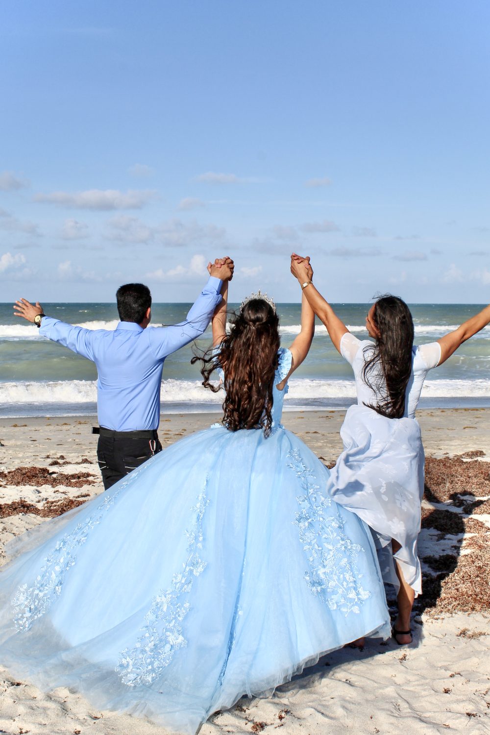Quinceañera in a sky-blue gown and two family members raising their arms in celebration at the beach