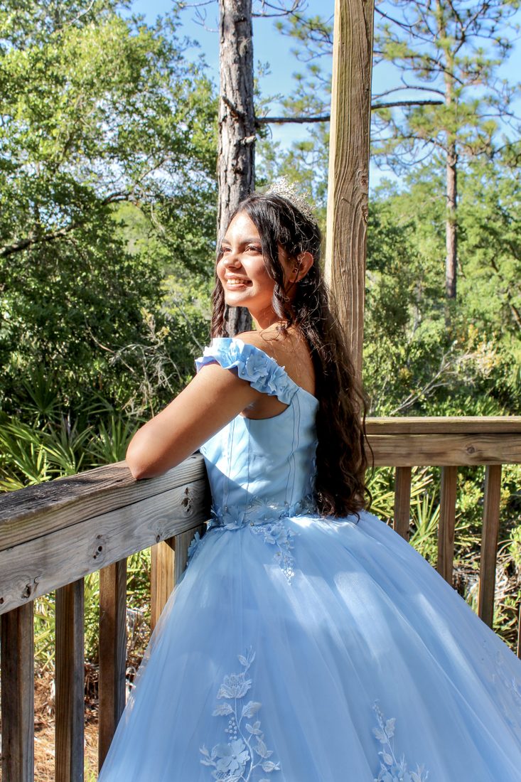 Girl in a sky-blue quinceañera gown smiling over her shoulder as she leans on a wooden railing among trees