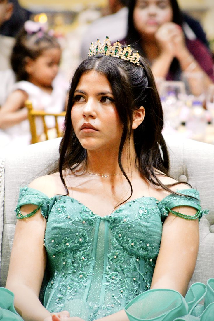 Girl in a mint-green gown and tiara seated in a white chair at her celebration, looking off to the side