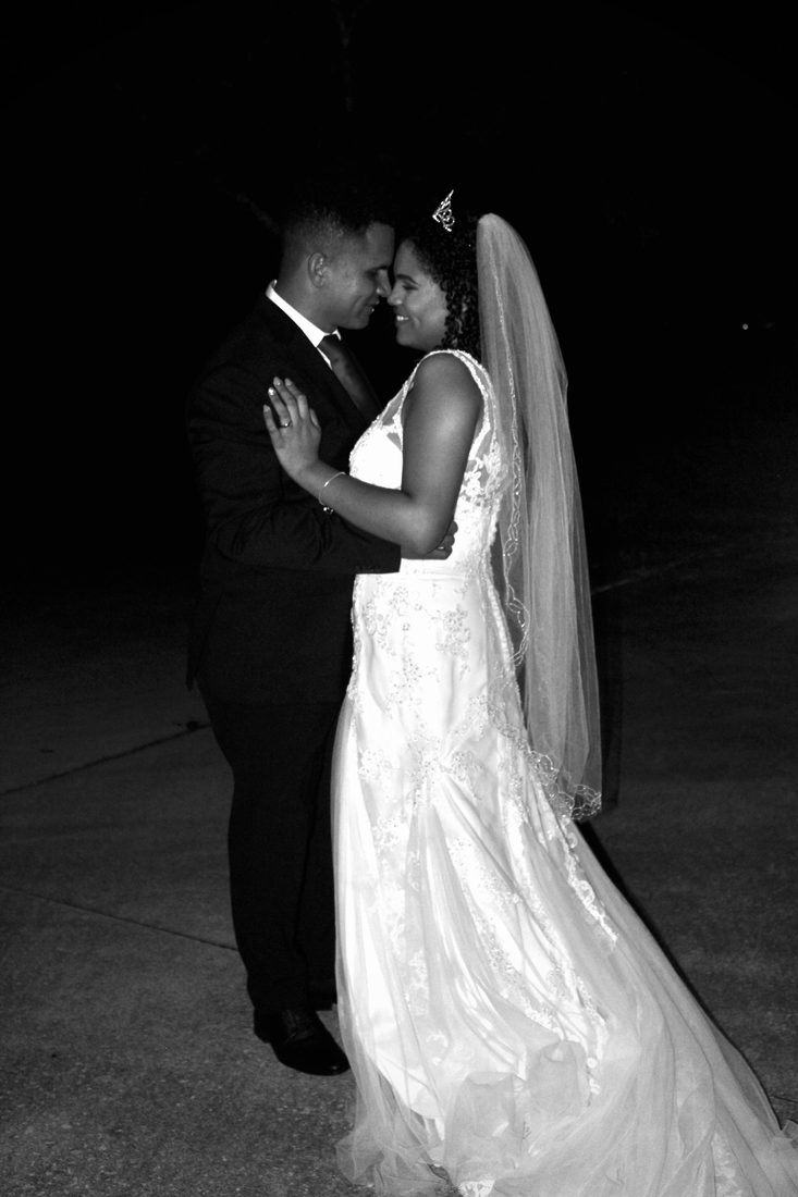 Black-and-white photo of a bride and groom slow dancing at night, her veil trailing behind her