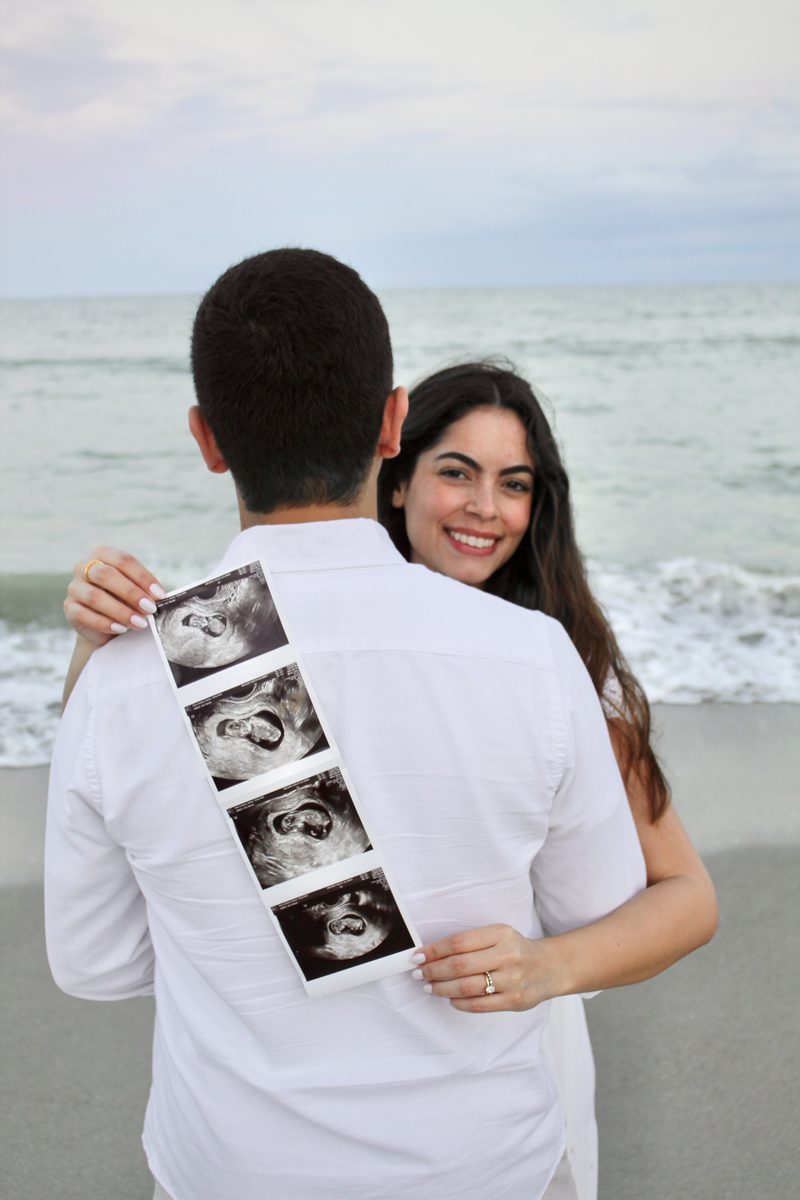 Couple embracing on the beach while he holds ultrasound pictures against his back