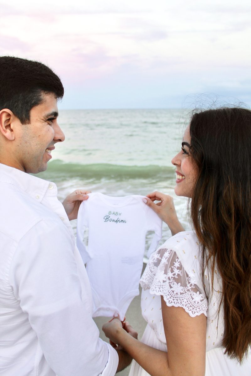 Couple on the beach at sunset holding up a baby onesie and smiling at each other