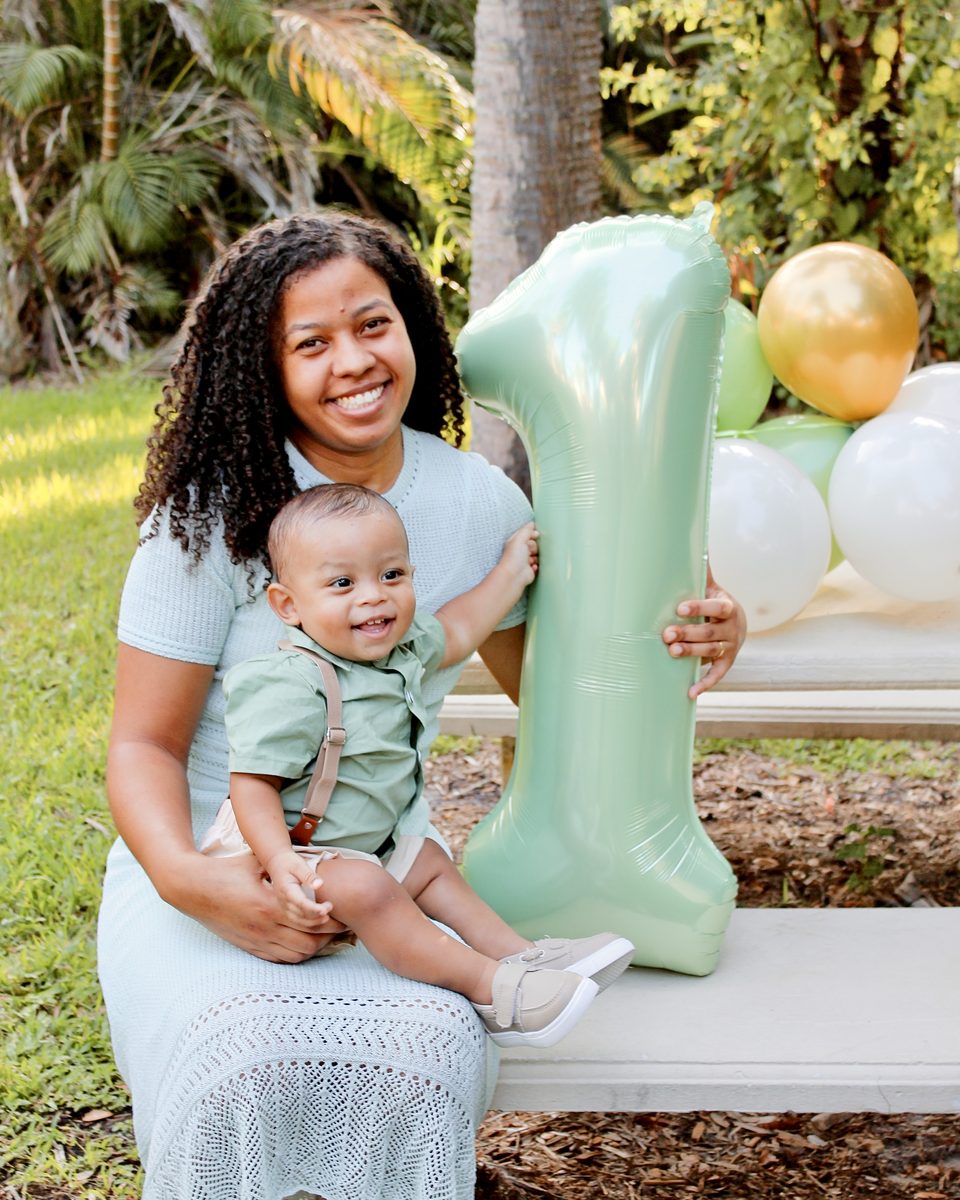 Mom in a mint dress holding her baby boy in front of a giant mint-green number 1 balloon