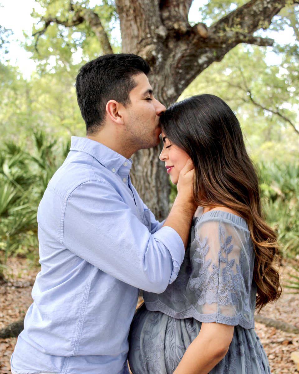 Expecting couple in soft blue outfits among trees, he kisses her forehead