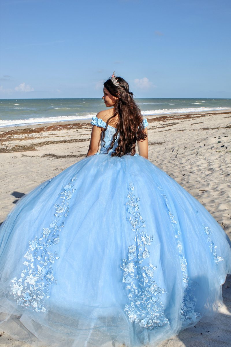 Girl in a sky-blue quinceañera gown seen from behind on the beach, looking out at the ocean