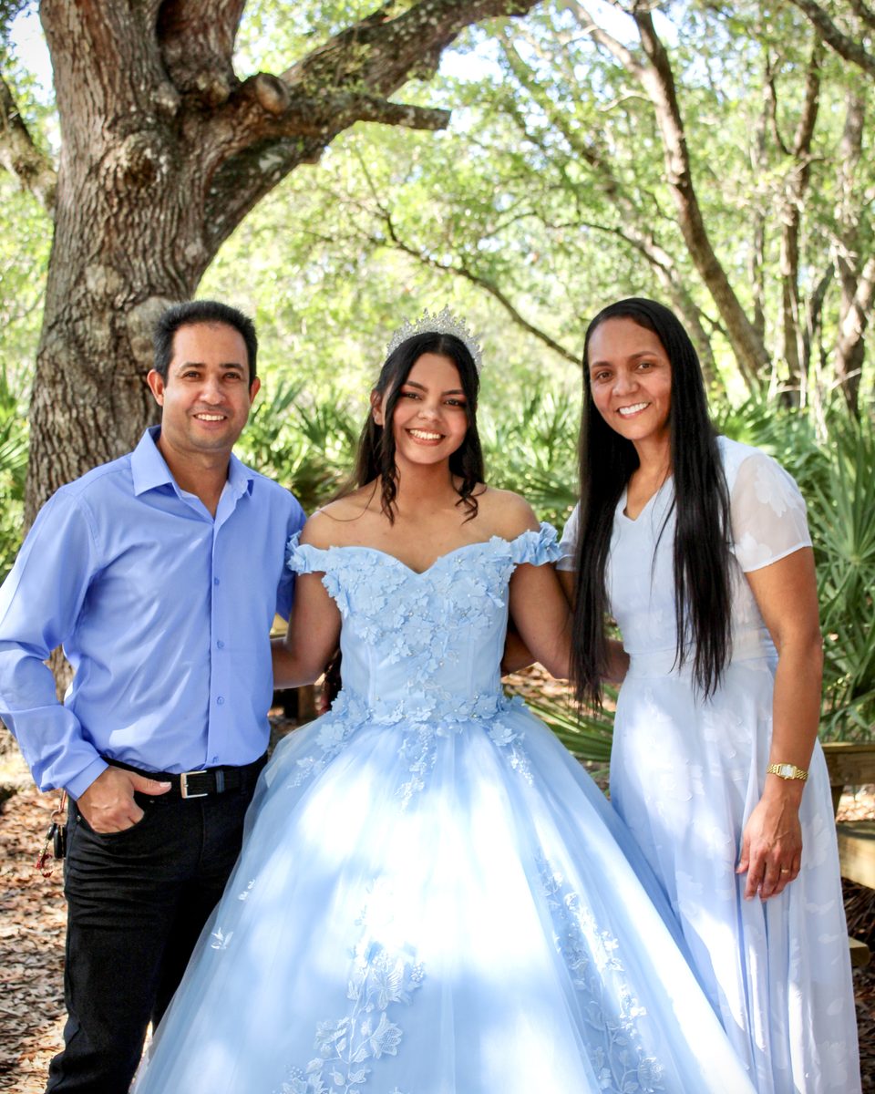 Quinceañera in a sky-blue gown posing with two family members under a big oak tree