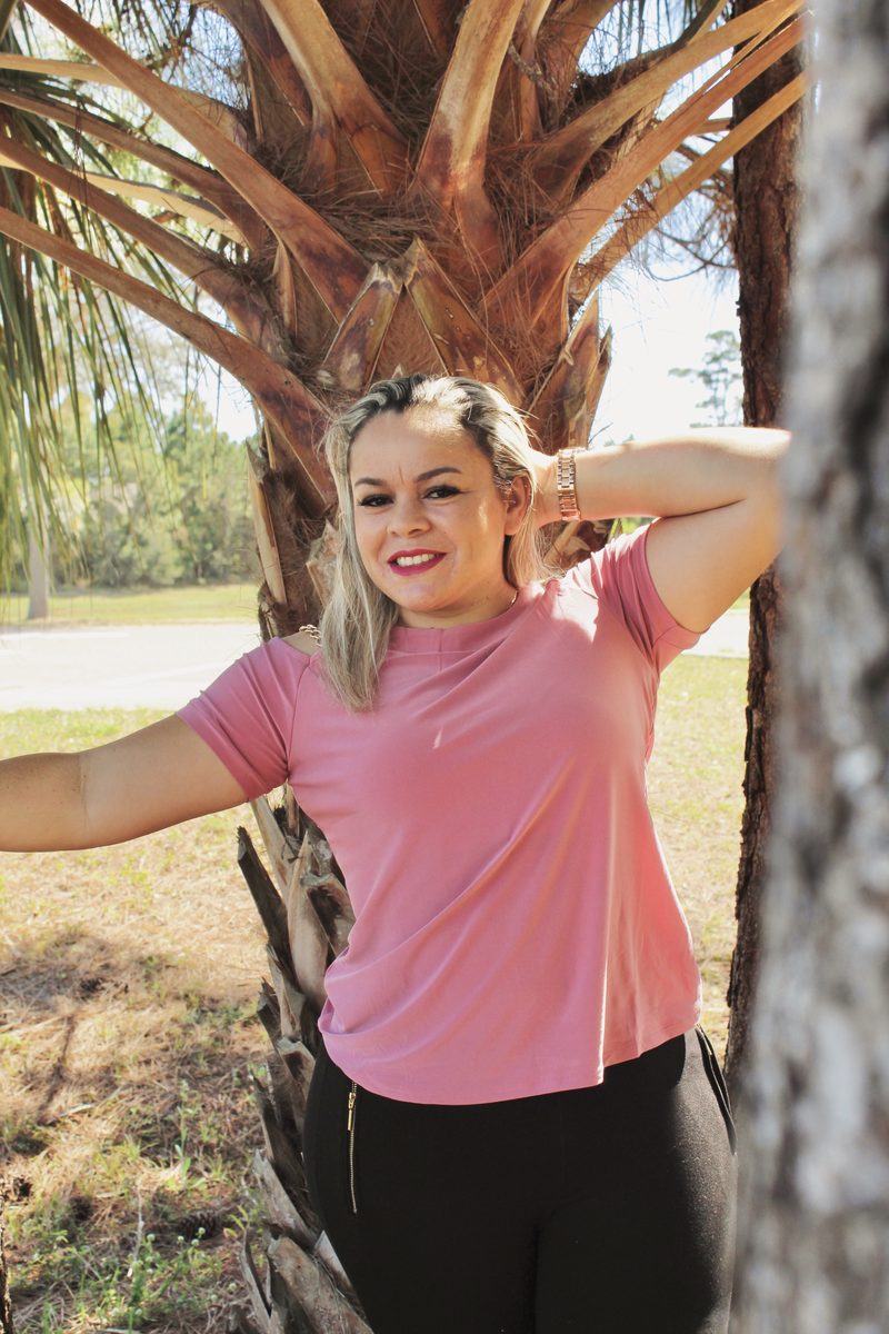 Woman in a pink top leaning against a palm trunk with one hand on her head, smiling