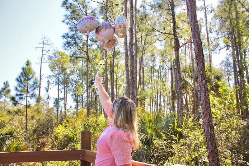 Woman in a pink top on a wooden deck among tall pine trees, holding up a bunch of balloons