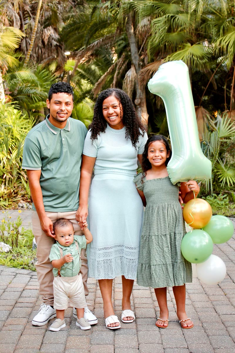 Family of four in matching green outfits among palm trees, holding a giant mint-green number 1 balloon and a bunch of balloons