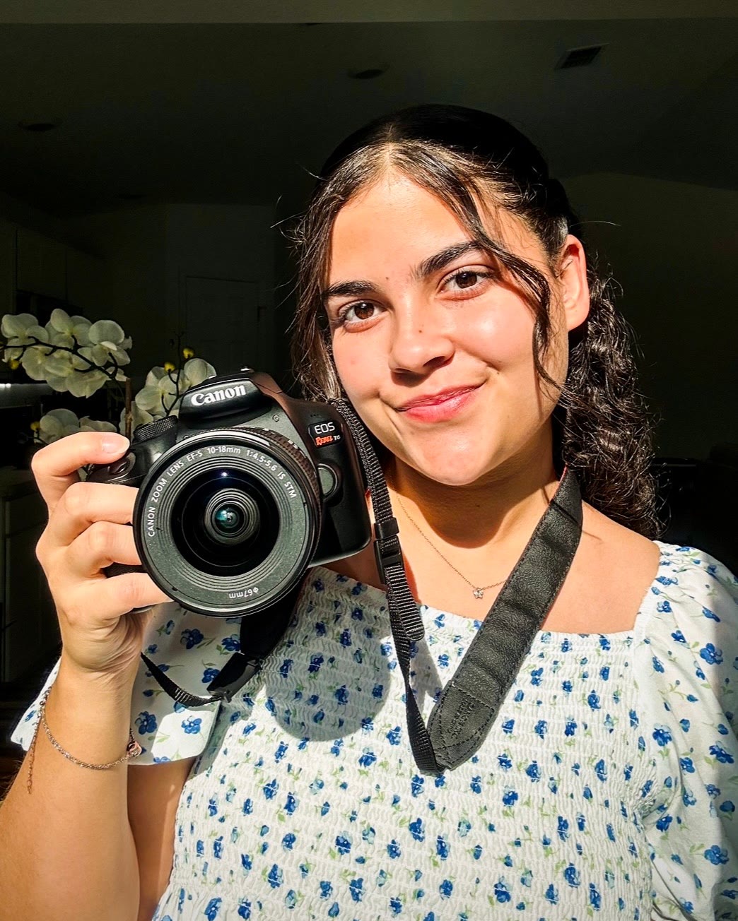 Kathlyn smiling in warm sunlight, holding a Canon camera and wearing a blue floral top