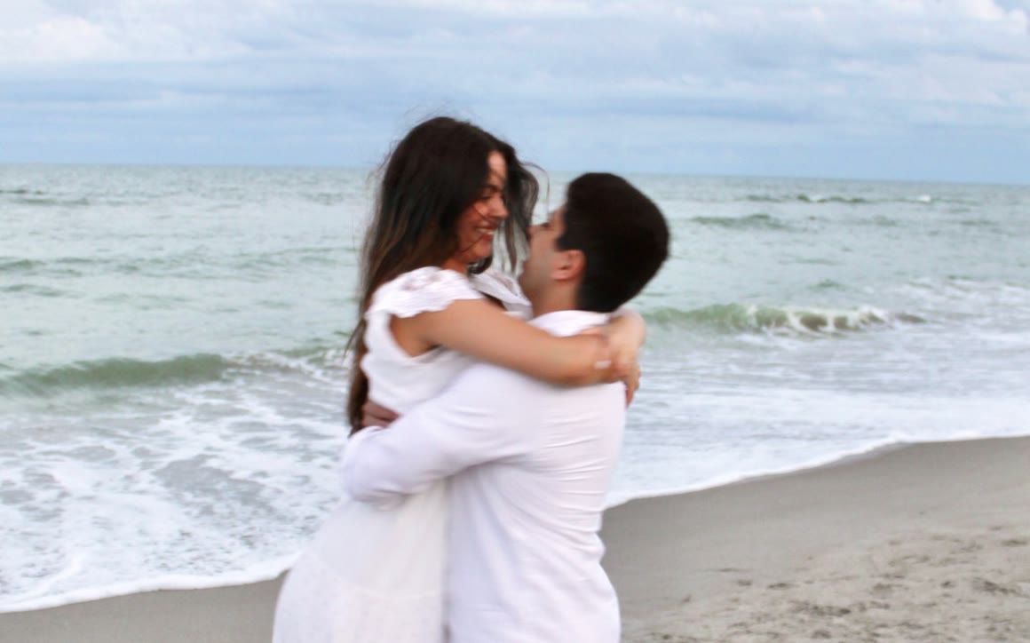 A couple in white embracing on a quiet beach at dusk as he lifts her off the sand, both laughing, with the ocean and soft clouds behind them