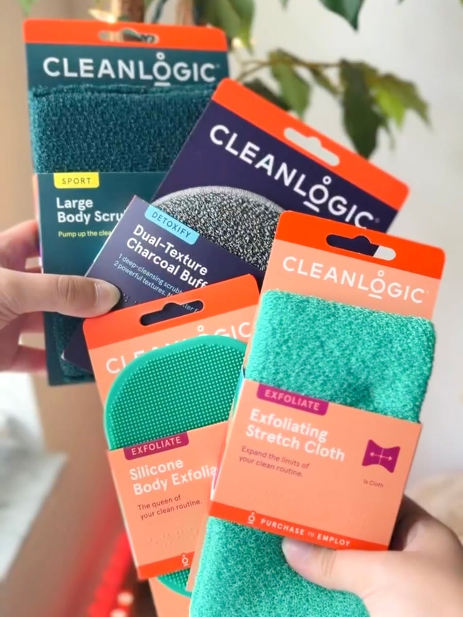 A fan of CleanLogic body scrubs, a charcoal buff, a silicone exfoliator and a stretch cloth held up in front of a plant